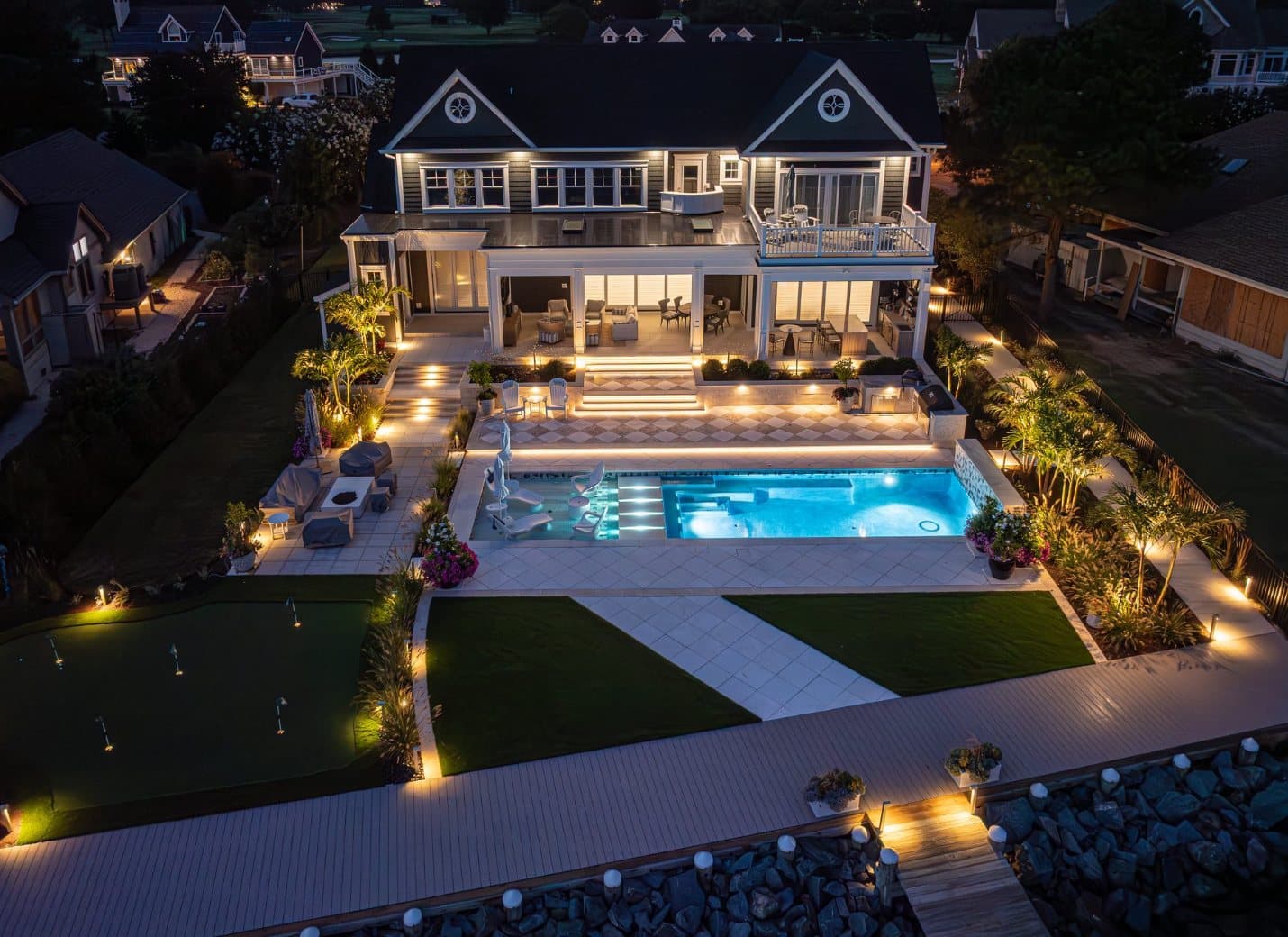 Aerial dusk view of a coastal home with pool, deck, path, and landscape lighting aglow
