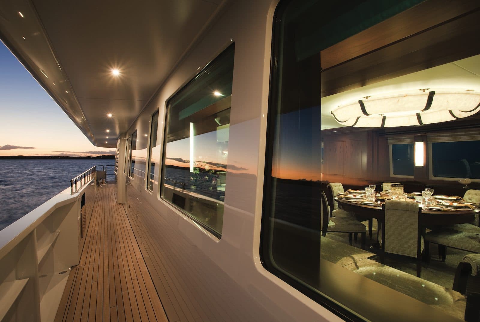 Yacht side deck at sunset, warm salon and set dining table glowing through the windows